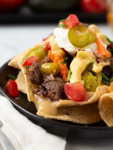 A close-up of a small black plate loaded with cheesy nachos sitting on a white marble countertop with a napkin and fork beside the plate.