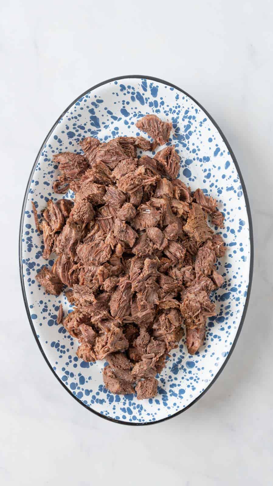 A blue and white speckled platter holding pot roast, chopped into bite sized pieces resting on a white marble countertop.