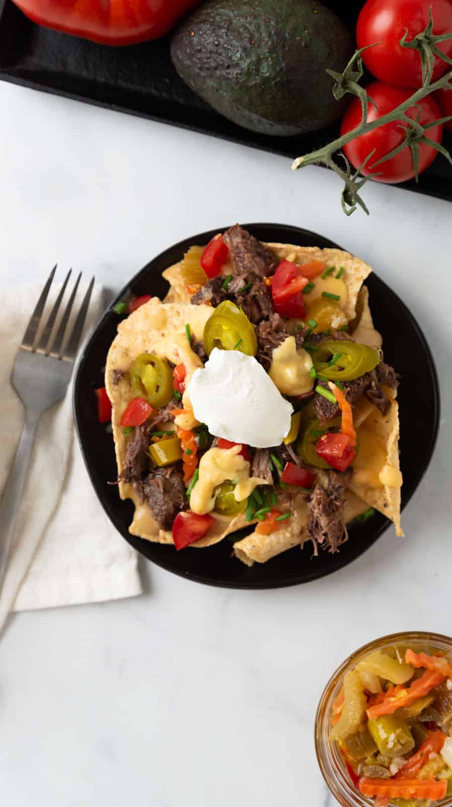 An overhead shot of a small black plate filled with pot roast nachos. A jar of giardiniera and tomatoes and avocados on the white marble countertop. A fork and cream linen napkin beside the plate.