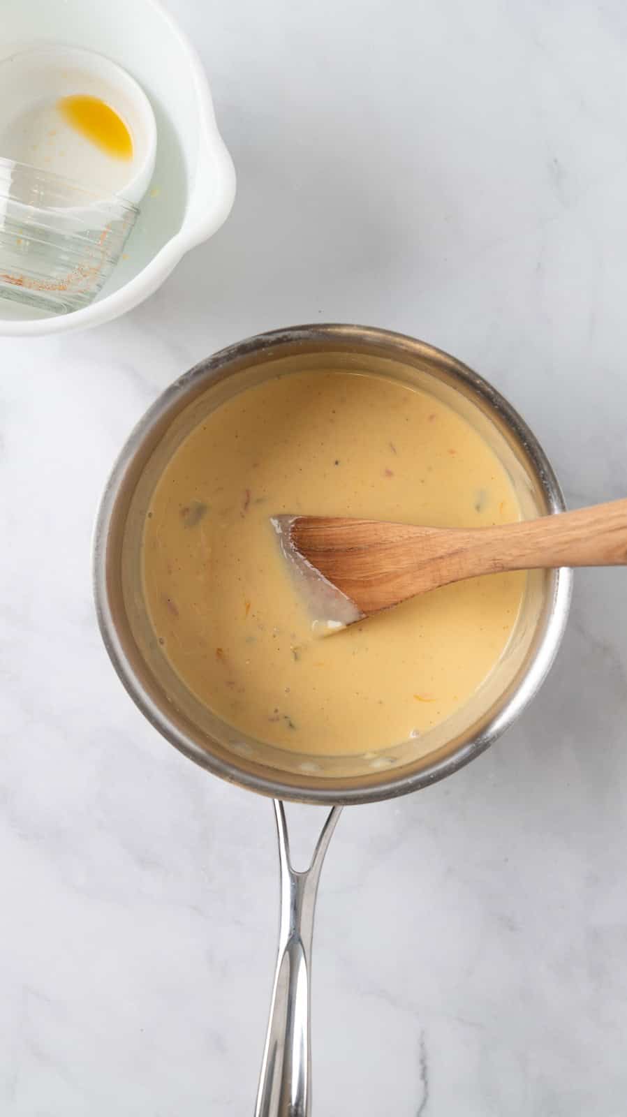 Giardiniera cheese sauce stirred together in a sauce pan with a wooden spoon. Empty ingredient bowls sitting near it on a white marble countertop.