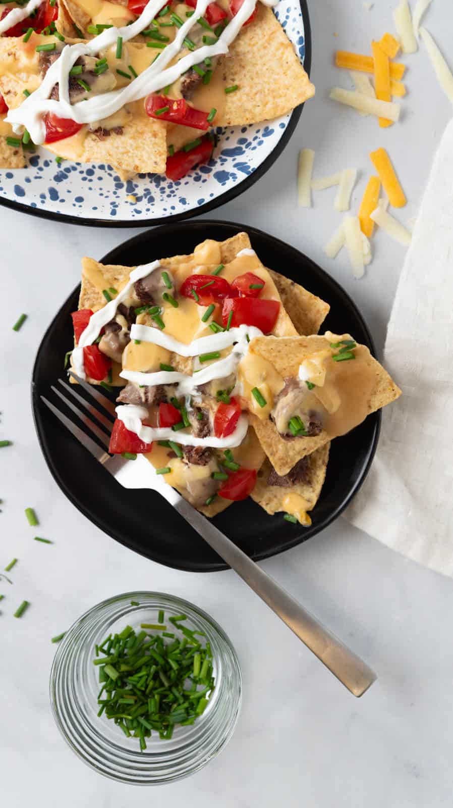 A small black plate of bright and colorful nachos with a fork. Part of the loaded platter close to the plate. Shredded cheese sprinkled on the white marble countertop. A small glass bowl holding chopped chives below the plate.