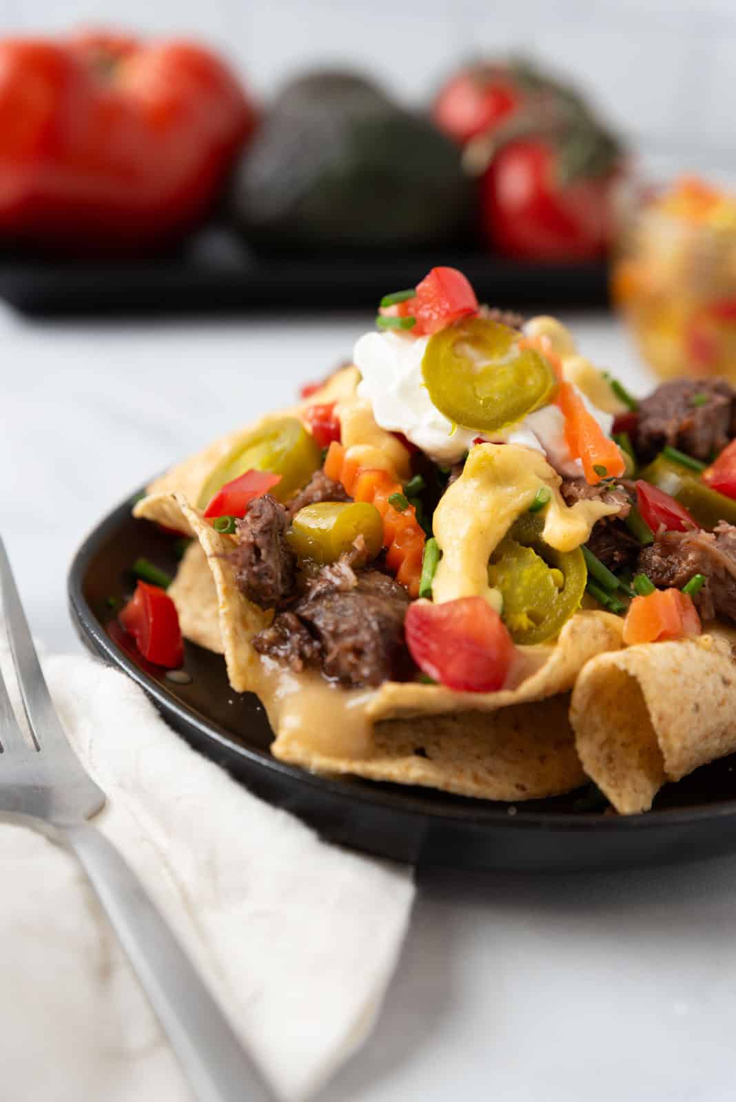 A close-up of a small black plate loaded with cheesy nachos sitting on a white marble coutnertop with a napkin and fork beside the plate.